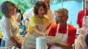 Group of adults in aprons gathered around a table in a bright kitchen, a man in red reads a recipe while others watch.