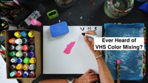 Top-down view of an art desk with paint bottles, brushes, and a hand writing on a white sheet; bold overlay reads 'Ever Heard of VHS Color Mixing?'