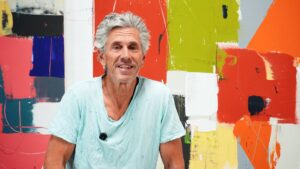 Smiling older man with gray hair in a light teal shirt poses in front of a bright abstract mural.