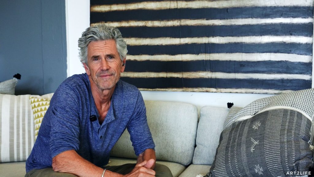 Older man with gray hair in a blue shirt sits on a light sofa, a lavalier mic clipped to his chest, in a living room with striped wall art behind him to the right.