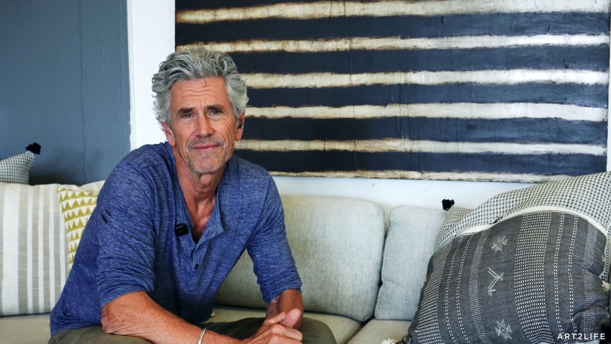 Older man with gray hair in a blue shirt sits on a light sofa, a lavalier mic clipped to his chest, in a living room with striped wall art behind him to the right.