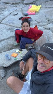 Two hikers resting on a rocky overlook with snacks, mugs, and gear scattered nearby. A yellow pad is in the background.
