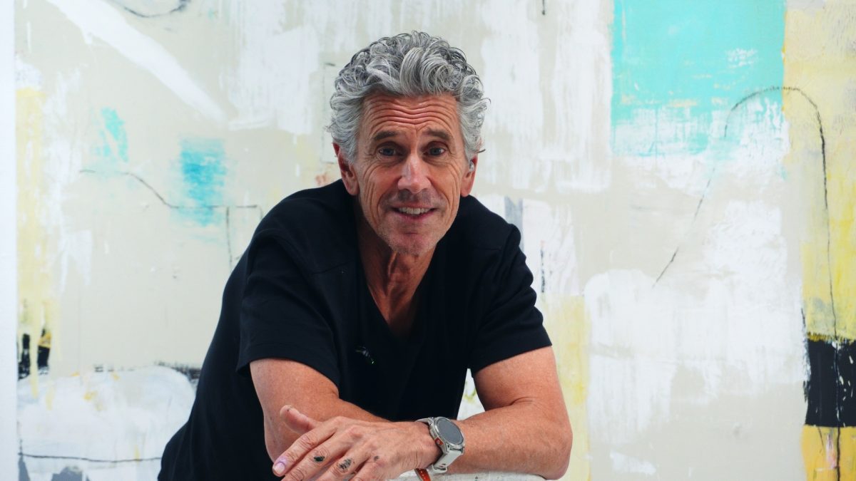 Smiling man with gray hair in a black shirt leaning on a painted wall backdrop with abstract art.