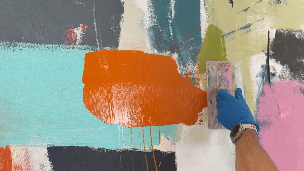 Gloved hand using a paint roller to apply orange paint on a colorful abstract mural with teal, pink, and gray sections.