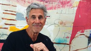 Older man with silver hair smiling at the camera in front of a vibrant abstract mural.