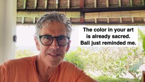 Self-portrait of a man in an orange shirt with round glasses, outdoors, next to a white sign that reads: 'The color in your art is already sacred. Bali just reminded me.'