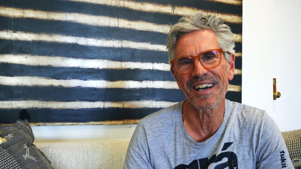 Smiling man with gray hair and orange glasses sits on a light sofa in front of a navy and white striped wall art.
