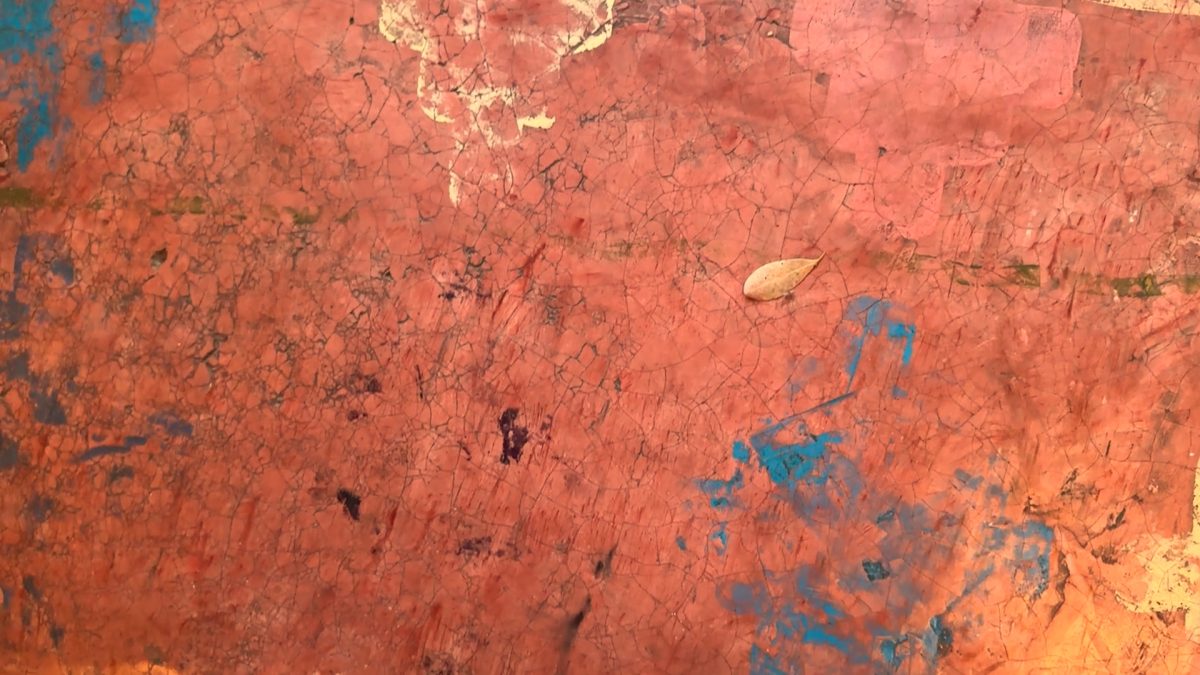 Cracked orange-paint surface with scattered blue marks and a single dried leaf near the center-right. (informative)