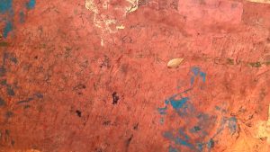 Cracked orange-paint surface with scattered blue marks and a single dried leaf near the center-right. (informative)