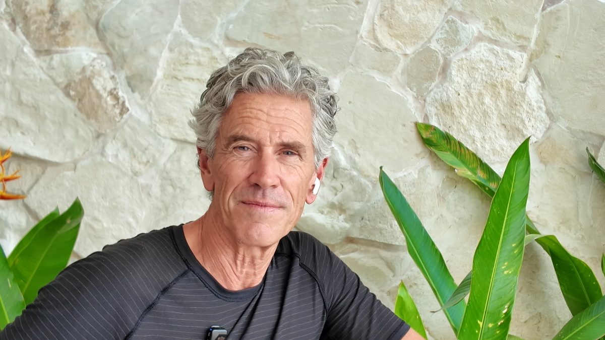 Portrait of a mature man with salt‑and‑pepper hair in a black striped shirt, sitting beside a stone wall with green tropical plants, wearing a wireless earbud in one ear.