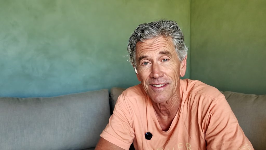 Smiling man with gray hair in a peach-colored shirt sitting on a beige couch against a green wall.