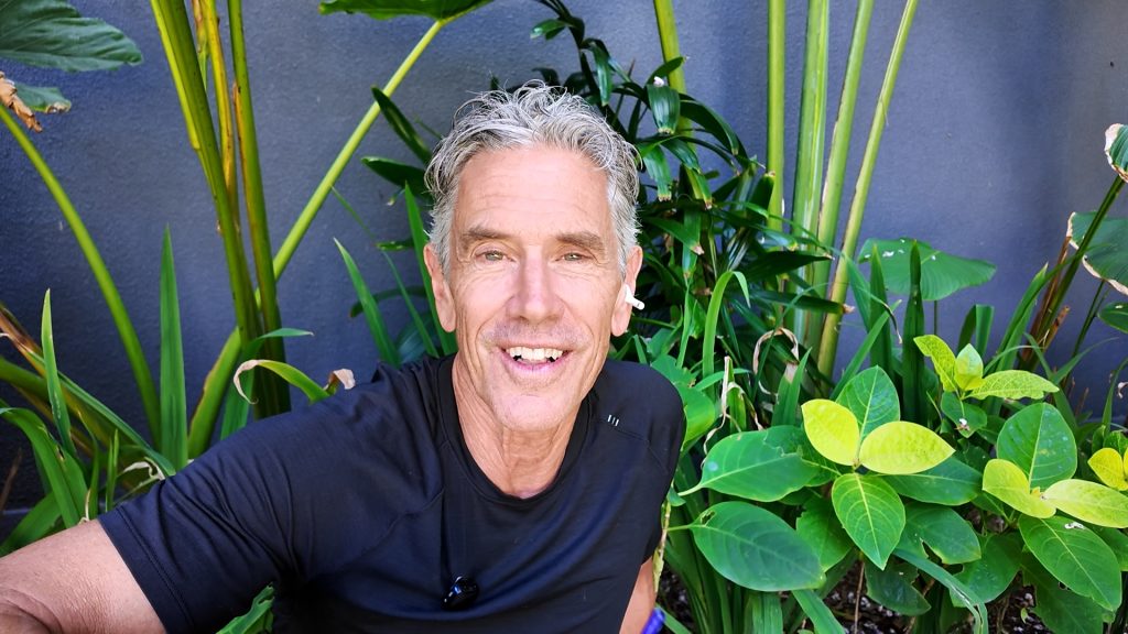 Smiling man in a black shirt taking a selfie among tall tropical plants against a dark wall behind him.