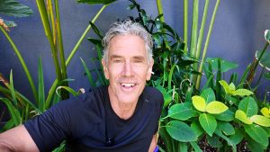 Smiling man in a black shirt taking a selfie among tall tropical plants against a dark wall behind him.