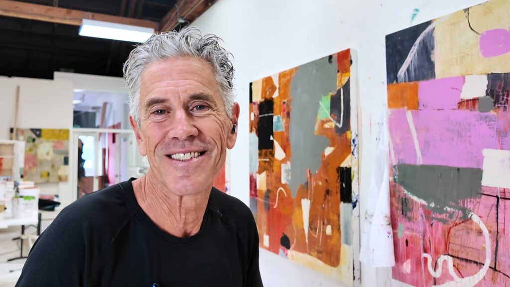 Smiling man in a black shirt stands beside a large colorful abstract painting in an art studio.